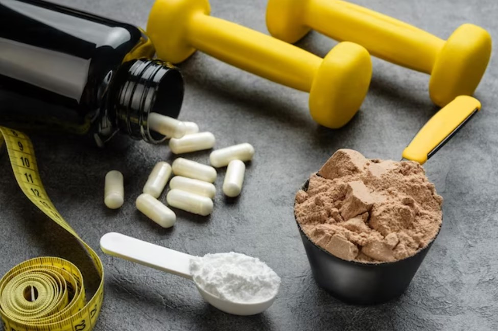 measuring spoons with chocolate protein, a jar with amino acid capsules, and yellow dumbbells