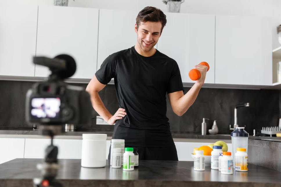 man in black shirt holding weights and filming on camera, bottles with pills and protein on the table