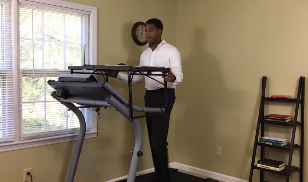 man in a white shirt and black jeans on a desk treadmill in a room